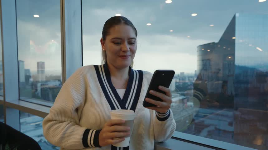 A young woman smiles while using her smartphone and drinking coffee by a window overlooking the city