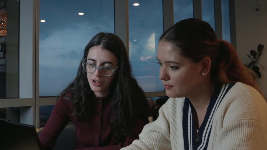 Two young professional women collaborate on a project on a laptop in a modern office with a view of the evening sky