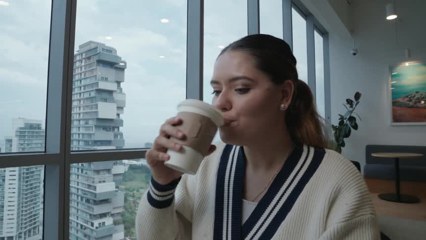 A smiling young woman works on her laptop and enjoys a coffee in a modern high-rise office with a city view in slow motion