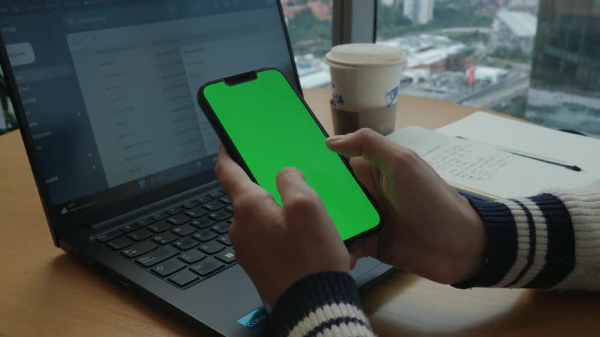A person scrolls through a green screen smartphone while sitting at a desk with a laptop and a city view in the background