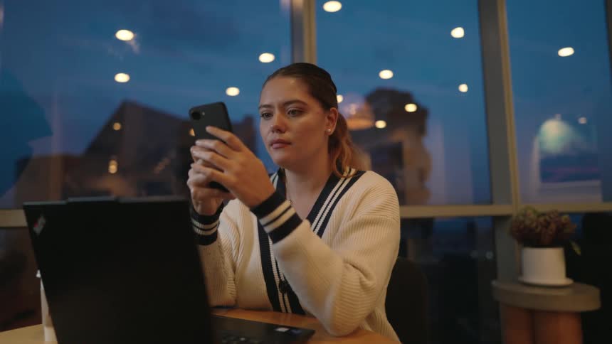A young professional woman uses her smartphone while sitting with her laptop in a modern office at night