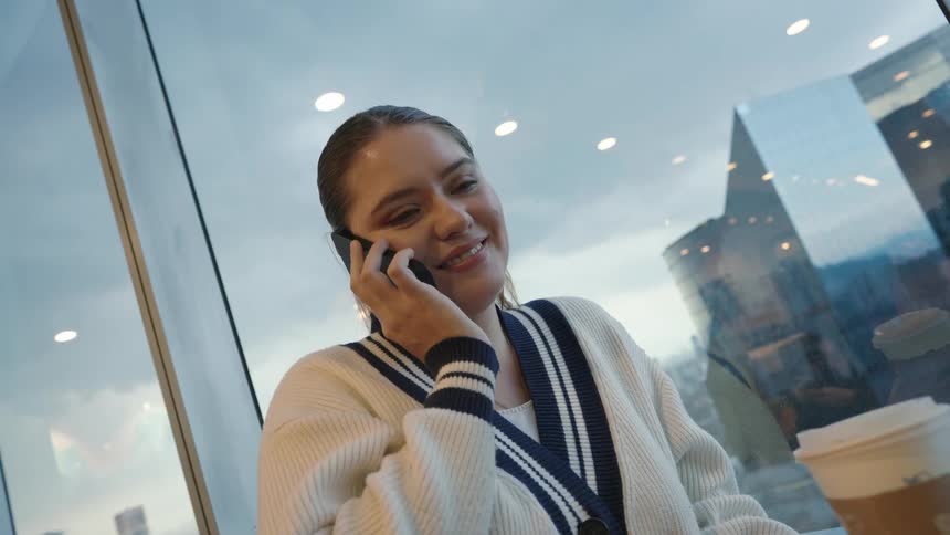 A young woman smiles while talking on her smartphone and holding a coffee cup next to a large window overlooking a modern cityscape