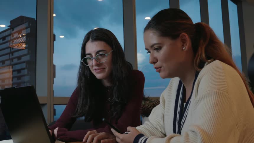 Two smiling young women work together on a laptop in a modern office with large windows overlooking the city at dusk