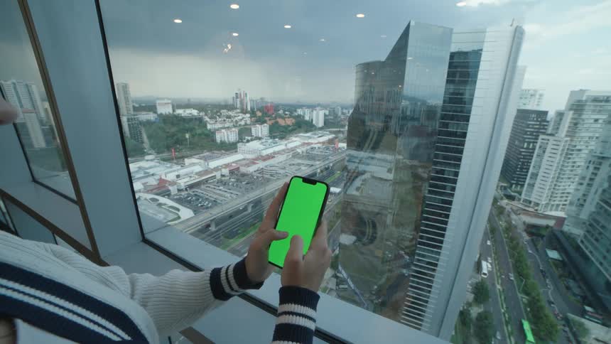 A person holds and interacts with a green screen smartphone inside a high-rise building overlooking a modern cityscape