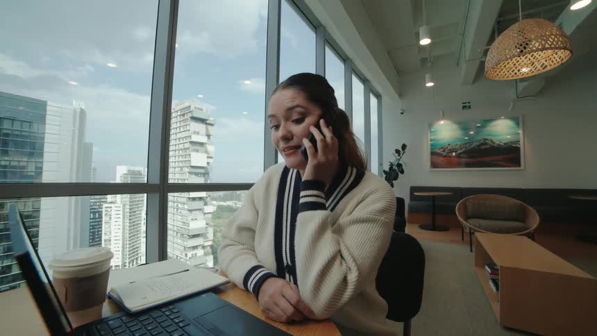 A happy businesswoman takes a phone call while working on her laptop in a modern high-rise office with a cityscape view