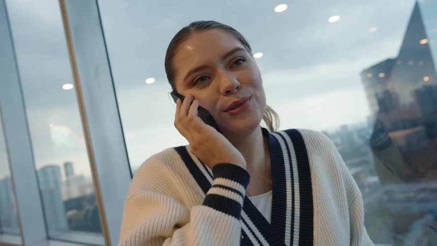 A happy young woman smiles while talking on her smartphone, enjoying coffee by the window of a modern office overlooking the city