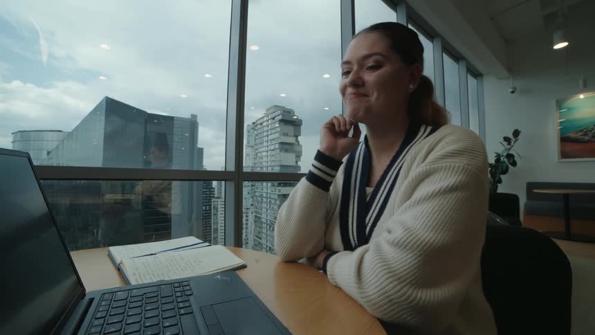 A young professional woman happily engages in a video call on her laptop in a modern office overlooking the city