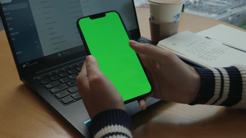 A person's hands hold and swipe on a vertical green screen smartphone while working at a desk with a laptop