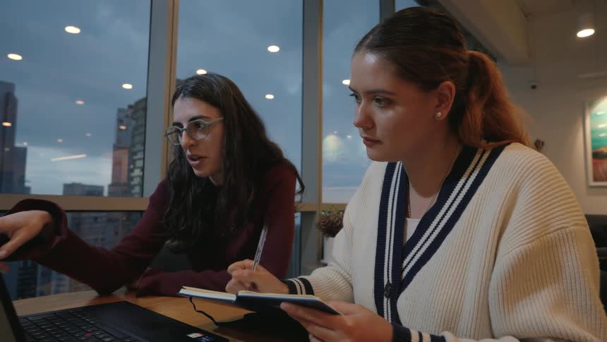 Two young women collaborate on a project, with one pointing at a laptop screen while the other takes notes in a modern office at dusk