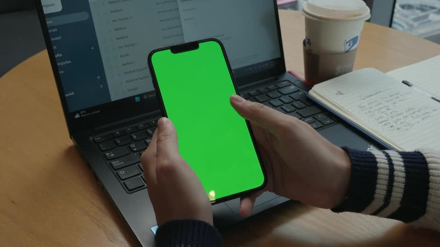 Close-up of a person scrolling on a smartphone with a green screen while sitting at a desk with a laptop and coffee