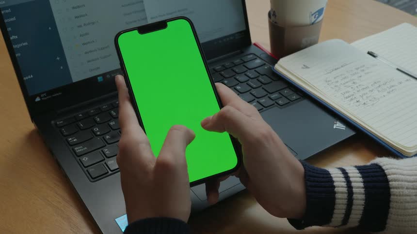 Close-up of a person's hands scrolling on a smartphone with a green screen at a desk with a laptop and notebook