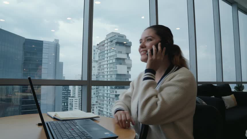 A young businesswoman works on her laptop and happily answers a phone call in her modern office overlooking the city skyline