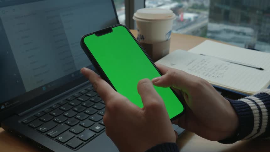 A person scrolls on a smartphone with a green screen while sitting at a desk with a laptop and coffee