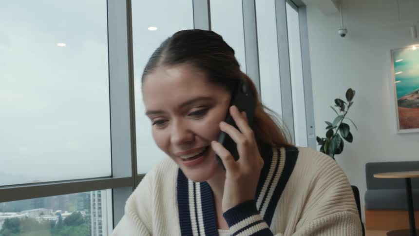 A close-up of an expressive young woman receiving exciting news on a phone call in a modern high-rise office