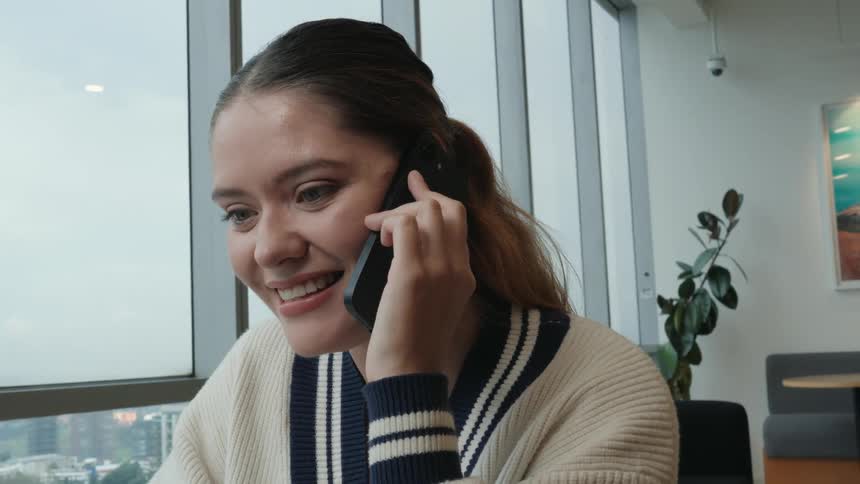 An excited young woman expresses surprise and happiness during a phone conversation by a large window