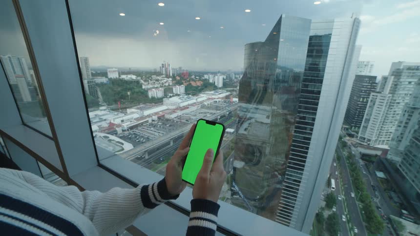A woman holds a smartphone with a green screen while sitting by a skyscraper window overlooking a bustling cityscape