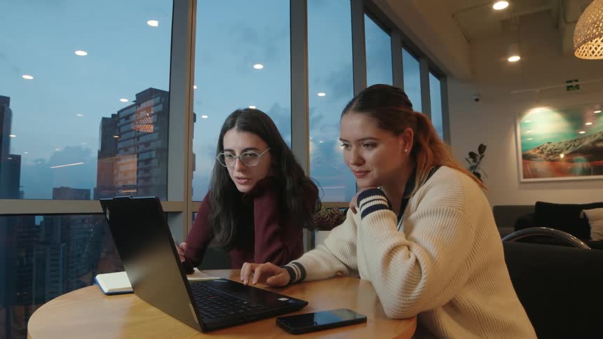 Two young women work together on a laptop, discussing and pointing at the screen in a modern office overlooking the city at twilight