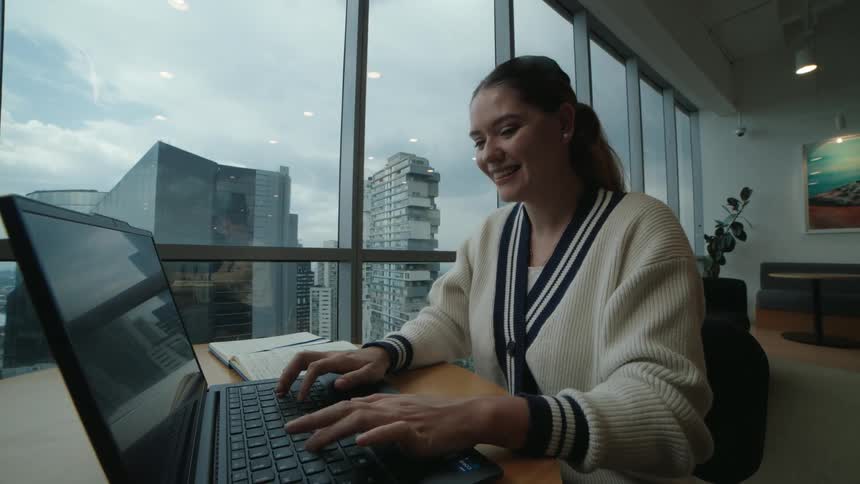 A smiling young businesswoman types on her laptop while sitting at a table in a modern office with a large window overlooking the cityscape