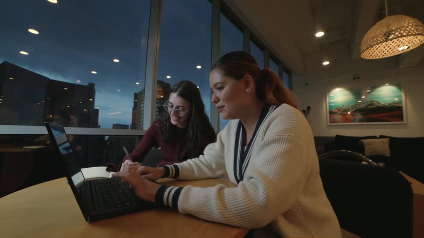 Two female colleagues collaborating on a laptop in a modern office with a city view at night