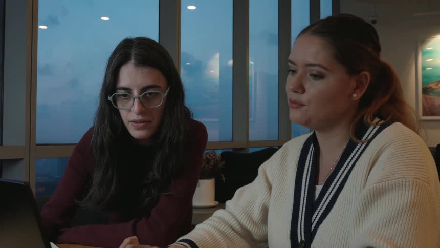Two female colleagues collaborate on a laptop in a modern office, with one yawning and laughing after a long day's work
