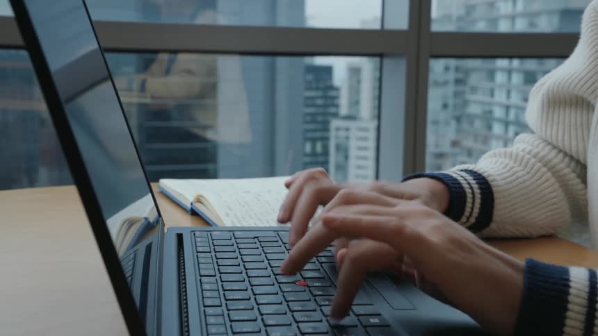 A close-up shot of a person's hands typing on a laptop keyboard at a desk in front of a window with a city view