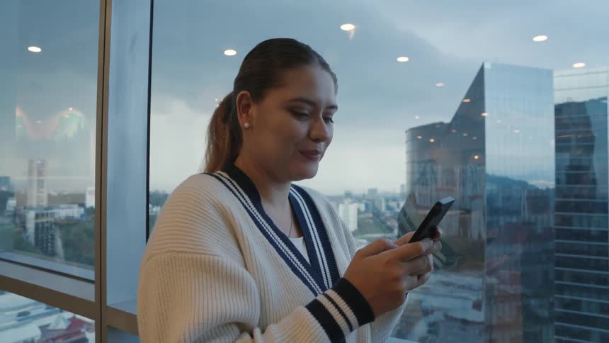 Happy young woman smiles while using her smartphone by a panoramic window with a city view in a modern high-rise