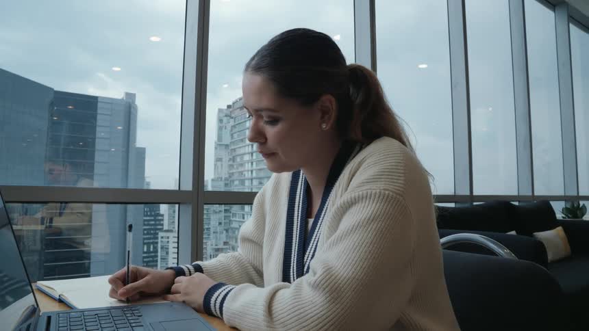 A thoughtful young woman writes notes in her planner before happily working on her laptop in a modern office with a city view