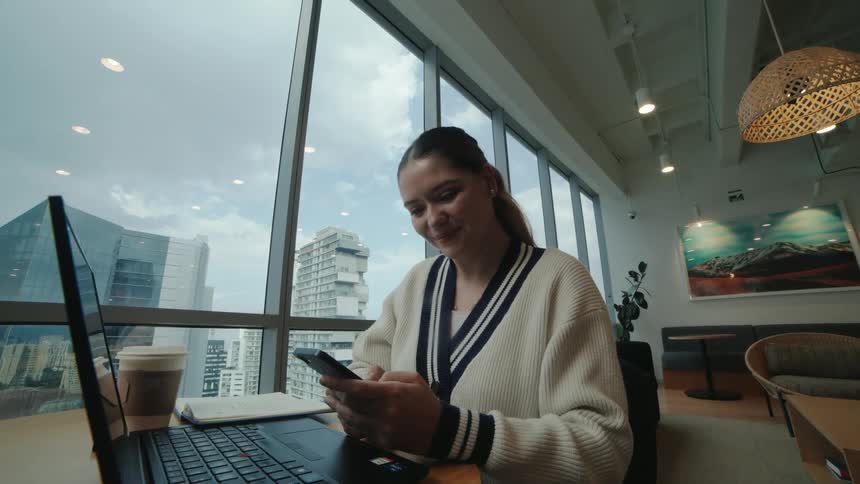 A happy young woman smiles while using her smartphone at her desk in a modern high-rise office with a city view