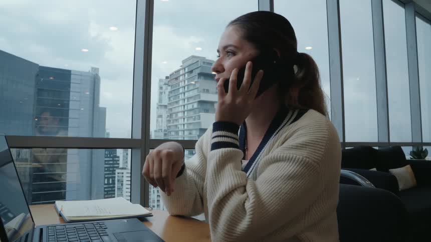 A young professional woman has an animated phone call in a modern high-rise office, her expression changing from serious to smiling