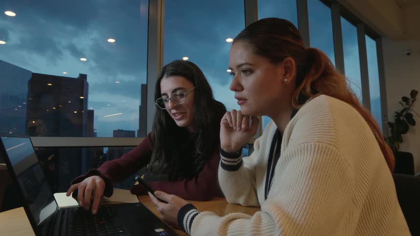 Two young women happily collaborate on a laptop in a modern office at dusk, pointing at the screen and laughing while working on a project