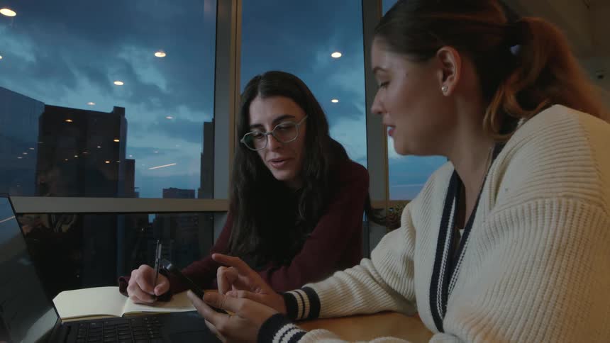 Two young female colleagues happily collaborate on a project in a modern office with a city view at dusk