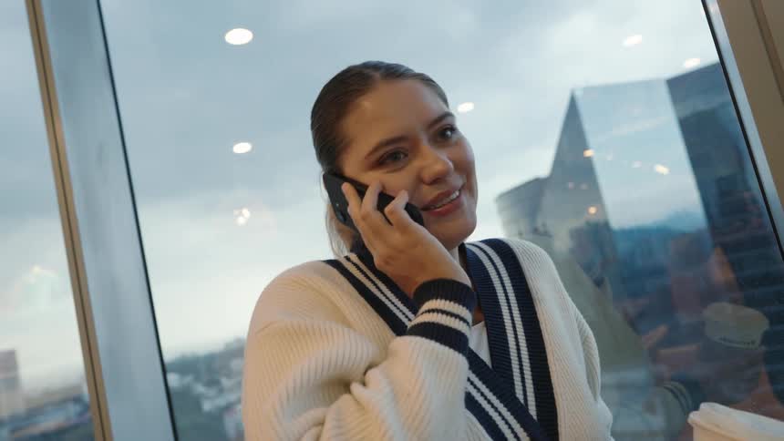 A young woman smiles while talking on her smartphone, holding a coffee cup by a large window overlooking a cityscape