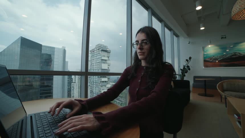 A focused young woman types on her laptop in a modern high-rise office with a sprawling cityscape view behind her