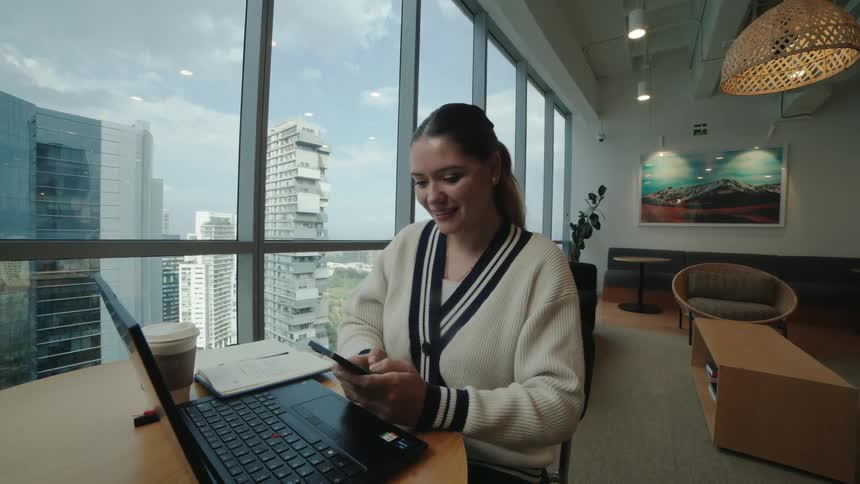 A happy young woman smiles while using her smartphone at a desk in a modern office with a city view