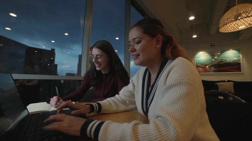 Two young women collaborate on a project in a modern office at night, one typing on a laptop while the other takes notes