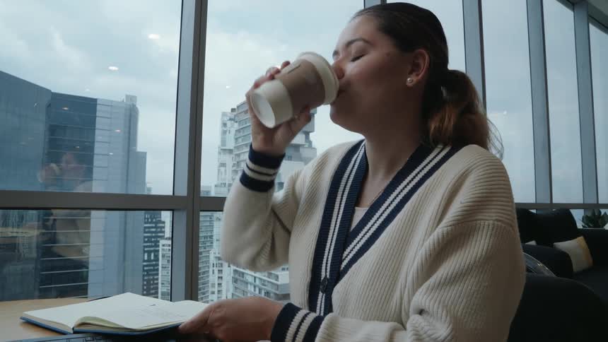 A professional woman enjoys a coffee break while writing in her notebook at a modern office with a city view