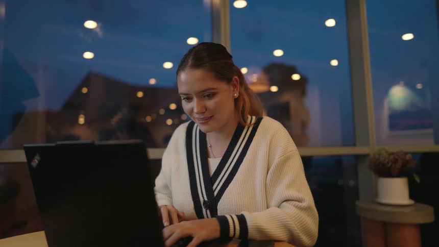 A focused young woman works diligently on her laptop in a modern office during the evening