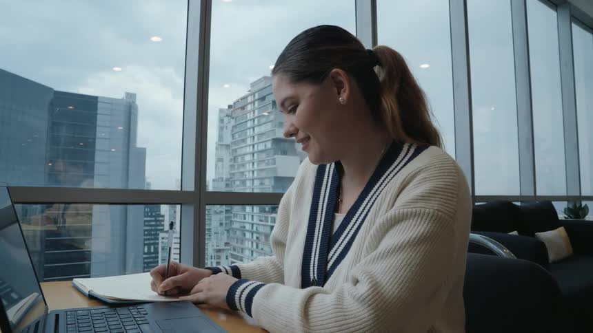 A young woman writes notes in her notebook before happily typing on her laptop in a modern office overlooking the city