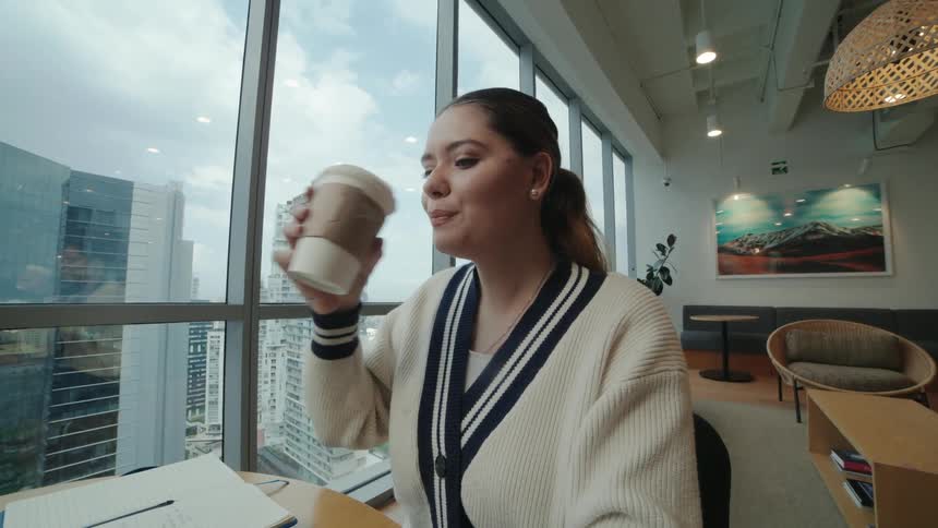 A young professional woman sips coffee while working on her laptop in a modern office with a beautiful city view, shown in slow motion