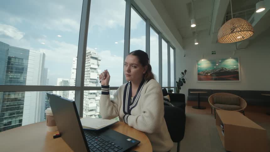 A thoughtful young businesswoman pauses to brainstorm ideas before writing them down in her notebook while working in a modern office with a city view