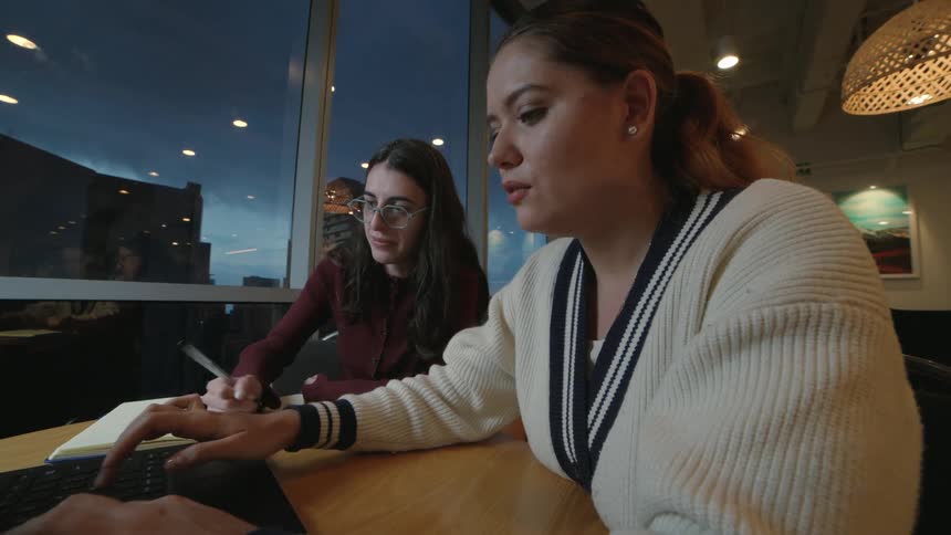 A close-up shot shows two young professional women collaborating on a laptop in an office with a city view at dusk