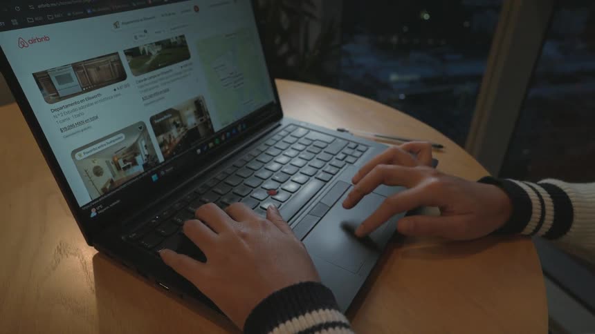 A person's hands browse and scroll through accommodation listings on the Airbnb website on a laptop at night