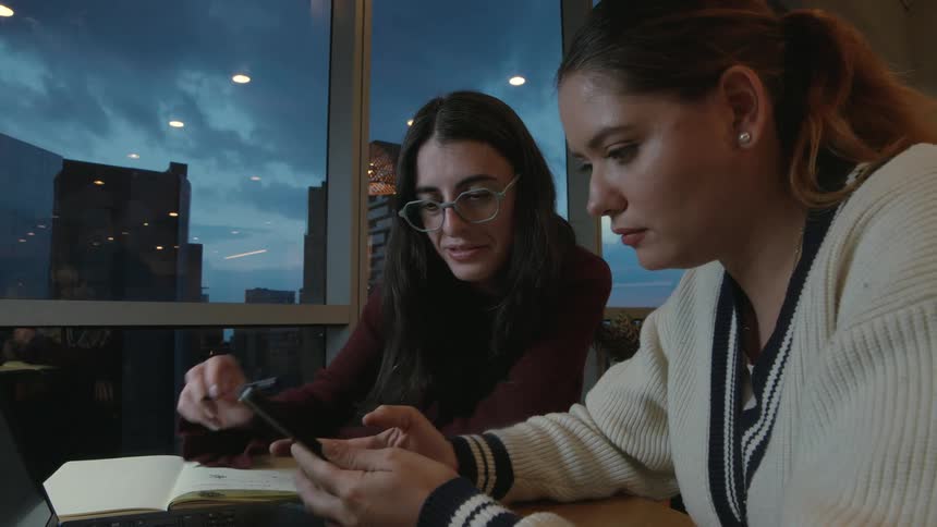 Two female students collaborate on a project using a smartphone and notebook in a cafe against a city skyline at dusk