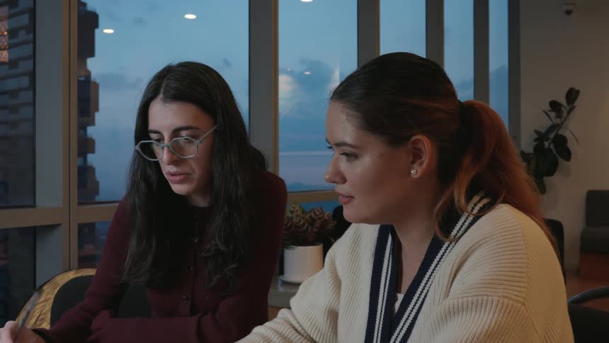 Two female coworkers discuss a project, with one taking notes, in a modern office with large windows overlooking the city at twilight