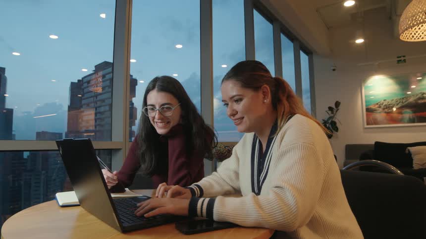 Two happy female colleagues collaborate on a project using a laptop in a modern office overlooking the city at dusk
