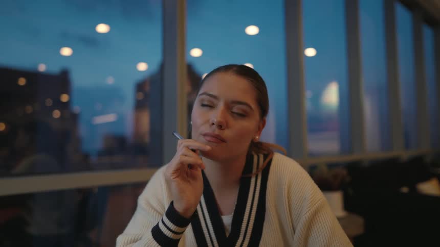 A thoughtful young woman holds a pen to her chin, deeply engrossed in her thoughts while sitting in a modern space at twilight