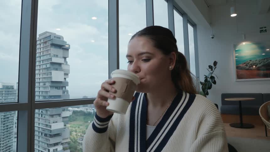 A smiling young woman works on her laptop and enjoys a cup of coffee in a modern office with a city view