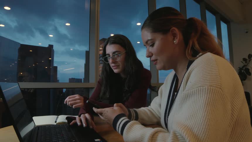 Two focused young women collaborate on a project, using a smartphone and laptop in a modern high-rise office as dusk settles over the city