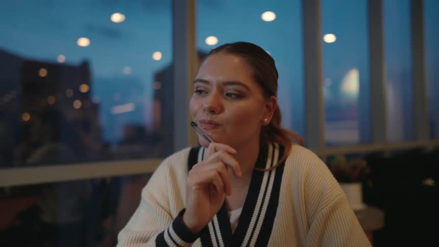 An inspired young woman thinks with a pen to her mouth, sitting by a window in a cafe during the evening