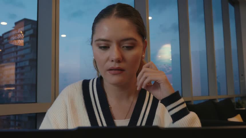 A young businesswoman opens her laptop in a city office at dusk and smiles at what she sees on the screen